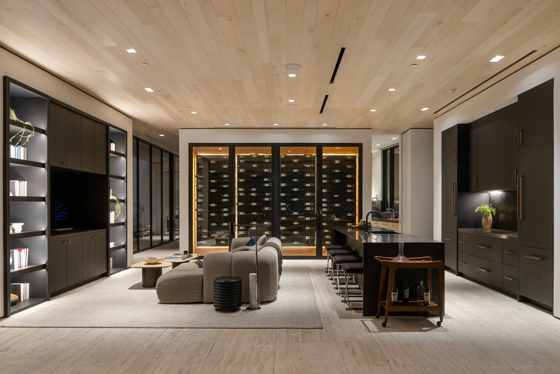Lounge with a pale wood plank ceiling, dark built-in shelving and an illuminated wine wall at the Laurel Way custom home