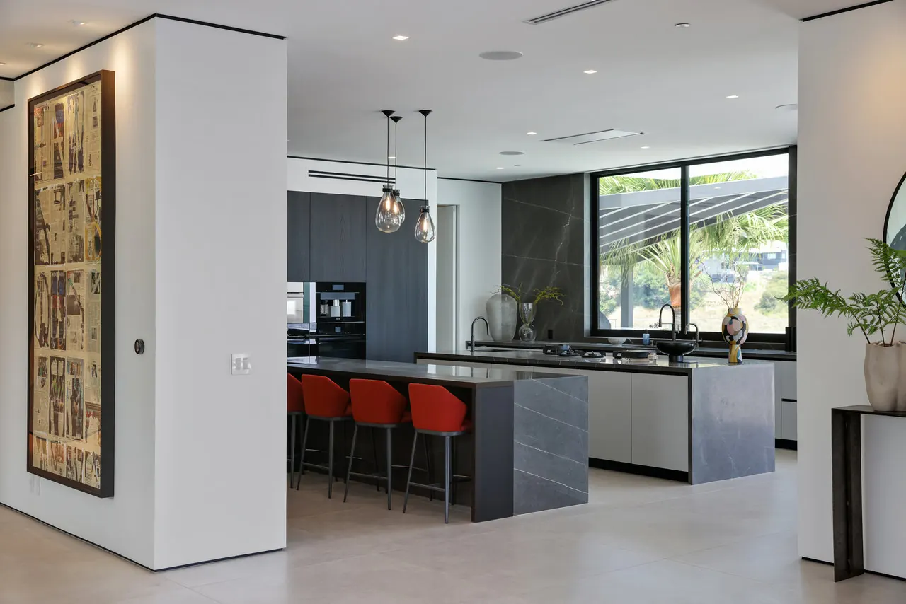 Kitchen with a dark stone island, red bar stools and glass pendants at the Laurel View Residence, a Hollywood Hills home built by Valley Vista Builders