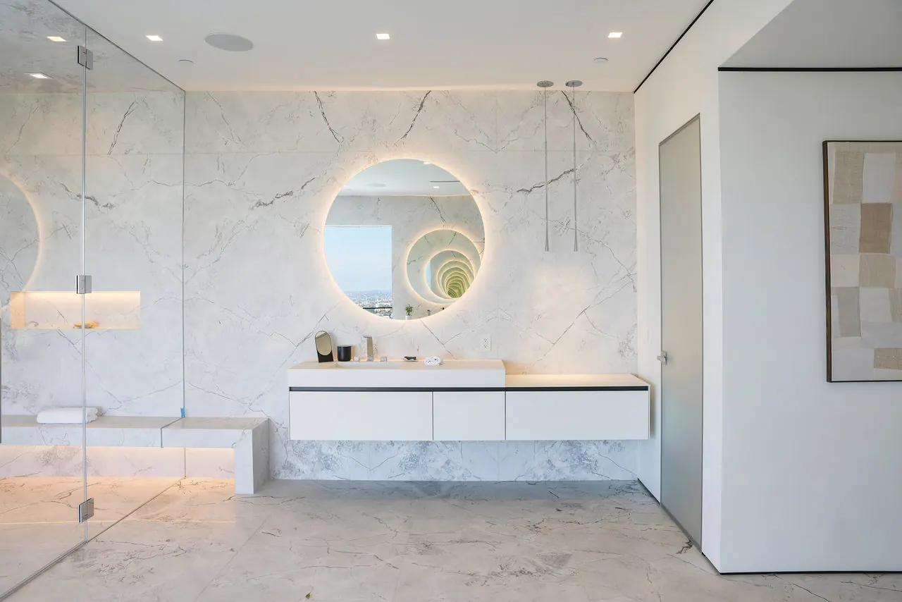 Bathroom with marble walls, a backlit round mirror and a floating vanity at the Laurel View Residence, a Hollywood Hills home built by Valley Vista Builders